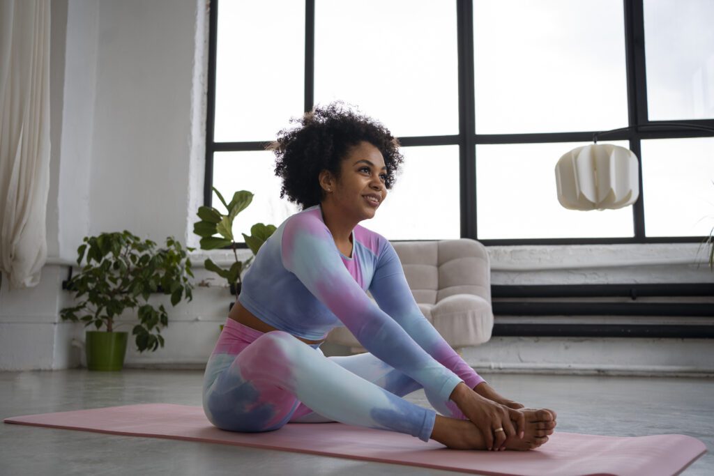 Mulher com roupa fitness colorida alonga-se em um tapete rosa, sorrindo em um ambiente iluminado com plantas e uma poltrona ao fundo